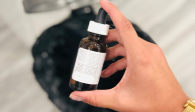 Hand holding a small amber glass dropper bottle with a white cap and label, containing a liquid product.